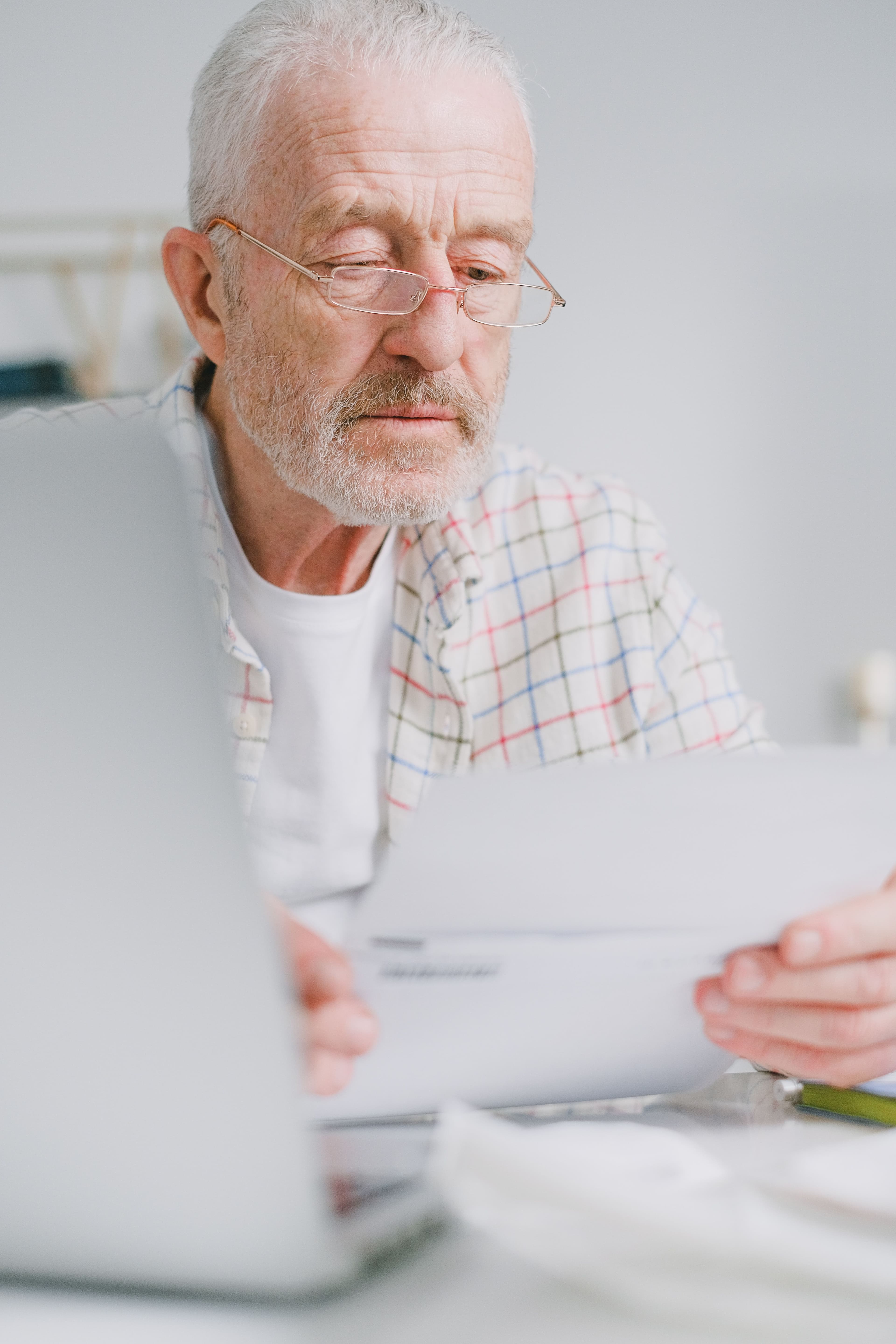 Homem idoso de óculos analisando documento com laptop ao fundo